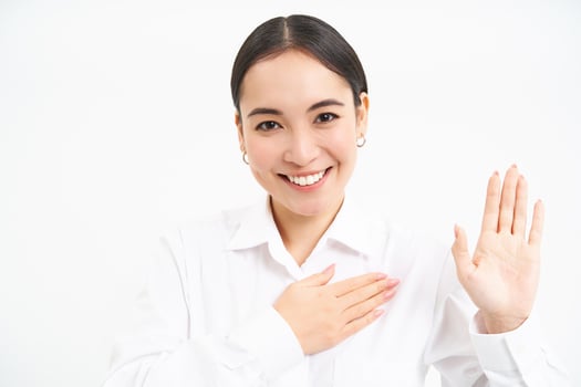 Friendly young businesswoman, asian lady raises one hand and puts palm on heart, makes promies, introduces herself, stands over white background photo