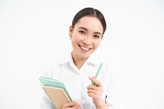 Beautiful confident korean woman, student with pen and notebooks, smiling and looking happy, standing over white background photo