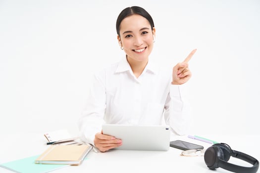 Portrait of young asian woman sitting in office, holding tablet and pointing at upper right corner, isolated on white background photo