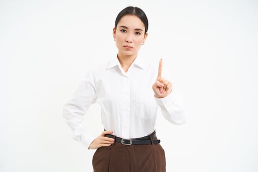 Confident young female manager, asian woman shows number one finger, explains smth, stands over white background photo