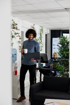 Arab project manager drinking coffee and planning strategy on laptop while standing near workplace table. Young professional holding portable computer and having tea in coworking space photo