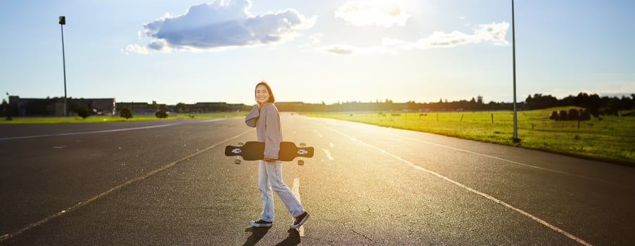 Young skater girl, teenager skating on cruiser, holding longboard and walking on concrete empty road photo