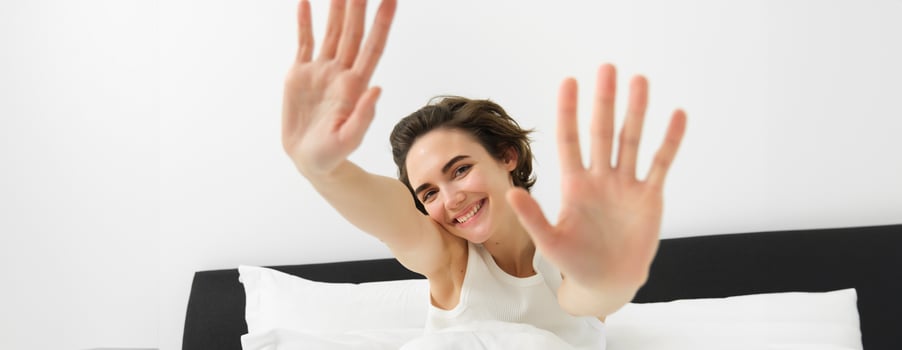 Beautiful young woman stretching her hands towards camera, waking up in morning, resting in her bed, smiling and looking happy photo