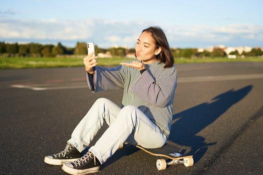 Happy asian girl sits on skateboard, takes selfie with longboard, makes cute faces, sunny day outdoors photo