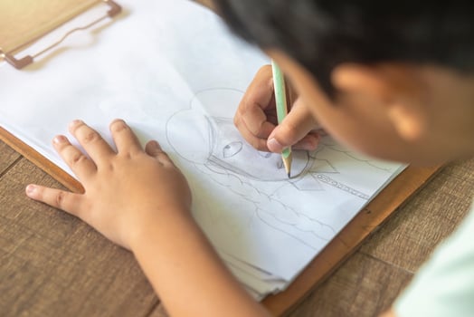 Close up portrait of Asian boy with black bangs, black eyes with a smiling face wearing a light green and lying on the floor of his house drawing a picture of a girl with braids. Education concept. photo