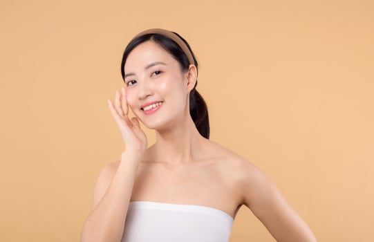 Beautiful girl asian model touching fresh glowing hydrated facial skin on beige background closeup. Beauty face young woman with natural makeup and healthy skin portrait. Skin care concept photo