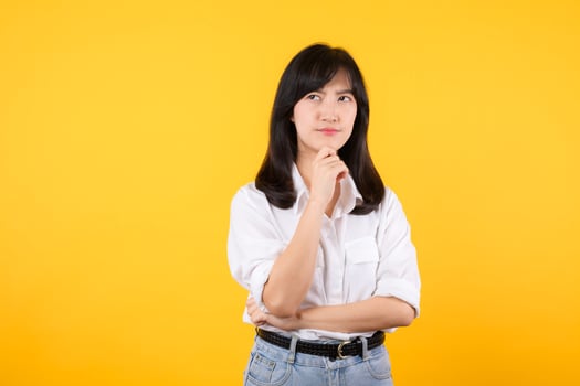 Thoughtful Asian woman keeps hand on chin looks pensively above wearing white shirt and jean plants poses against yellow background blank copy space for your advertising content thinks about future. photo