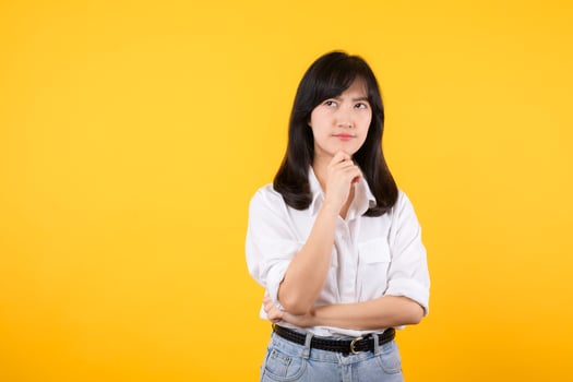 Thoughtful Asian woman keeps hand on chin looks pensively above wearing white shirt and jean plants poses against yellow background blank copy space for your advertising content thinks about future. photo