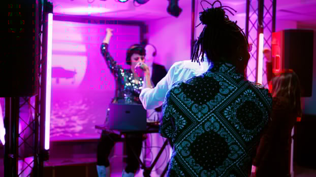 Young adult putting music at party, working as DJ at discotheque with friends. Woman using audio mixing station on stage with discolights, having fun with group of people. Handheld shot. photo