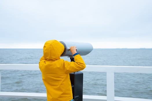 A child in a jacket stands on a white pier. looking through binoculars at the sea. Cold weather. back view photo