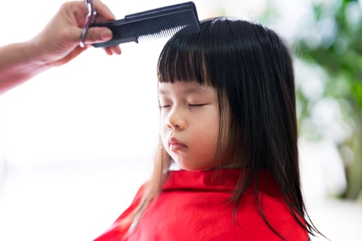 Asian kid girl sits still with her eyes closed while hairdresser is styling her hair in order to beautify it. 4 year old child is held in red veil to prevent hair from getting stuck in her clothes. photo
