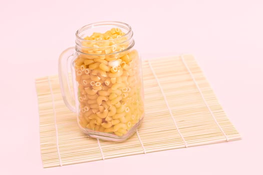 Uncooked Chifferi Rigati Pasta in Glass Jar on Bamboo Mat on Pink Background. Fat and Unhealthy Food. Scattered Classic Dry Macaroni. Italian Culture and Cuisine. Raw Pasta photo