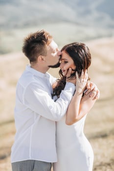 A stylish model couple in the mountains in the summer. A young boy and a girl in a white silk dress are walking on the slope against the background of the forest and mountain peaks. boy kisses a girl photo