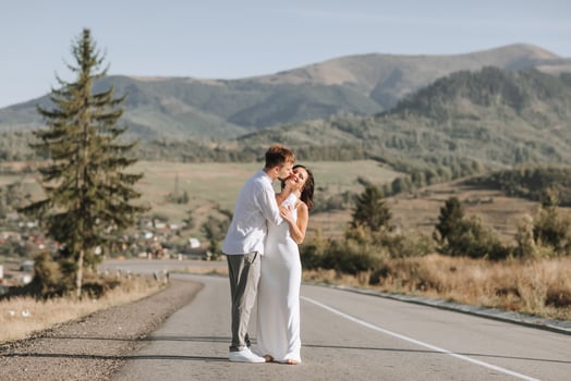 A stylish groom in a white shirt and a cute brunette bride in a white dress are walking on an asphalt road against the background of a forest and mountains. Wedding portrait of newlyweds. photo