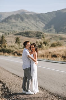 A stylish groom in a white shirt and a cute brunette bride in a white dress are hugging on the side of the road against the background of the forest and mountains. Wedding portrait of newlyweds. photo