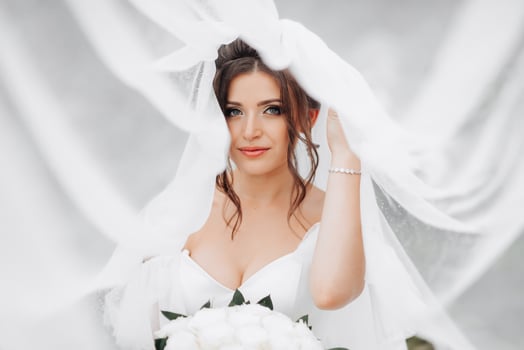 Curly brunette bride in a white dress, covered with a veil, poses for the camera with a bouquet of roses. Portrait of the bride. Beautiful makeup and hair. Wedding in nature photo