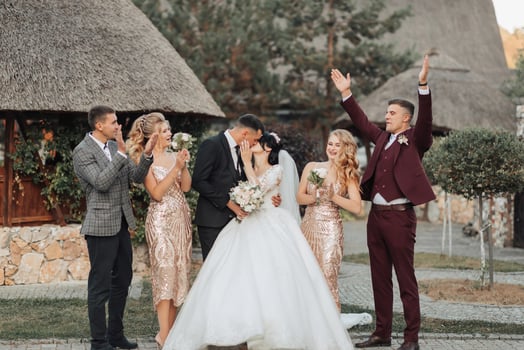 Front view of newlyweds standing and kissing, between friends, friends having fun. Girls in golden dresses hold bouquets, the bride in a voluminous dress, the groom and his friends in suits photo