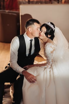 Wedding portrait of the bride and groom. Indoor photography with beautiful light. Stylish Groom in a black vest kisses a brunette bride on the cheek, sitting hugging indoors. Couple in love photo