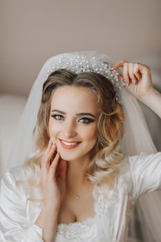 A blonde bride is wearing a satin robe and sitting in her room, posing and showing off her tiara and veil. Beautiful hair and make-up, open bust. Wedding portrait. Sincere smile photo