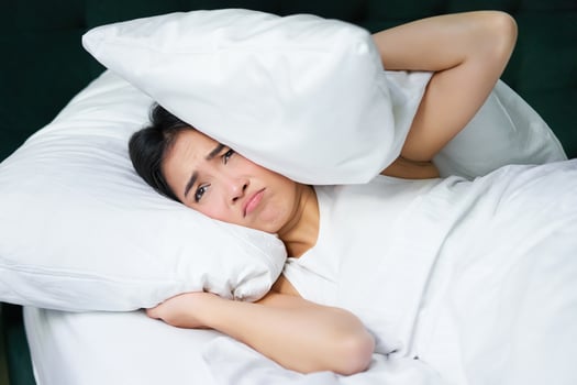 Sad girl with insomnia. Young asian woman lying in bed, cover her ears with pillow, cant sleep, partner snors, loud noise at night disturbing her photo
