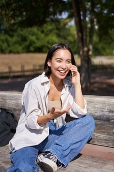 Portrait of happy young asian woman talking on mobile phone in park, sitting on bench and having a telephone call, chatting lively photo