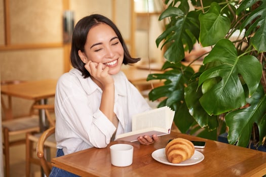 Romantic asian woman sitting with book in cafe, eating croissant and drinking coffee, reading and smiling, enjoying alone time photo