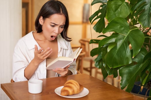 Portrait of asian woman looking shocked at book pages, reading something interesting, concentrating, sitting in cafe with coffee and croissant photo