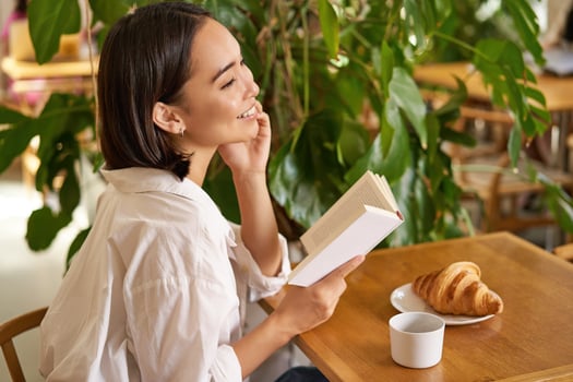 Beautiful young asian woman with a book in hands, sitting in cafe, drinking coffee and eating croissant, smiling, looking mysterious photo