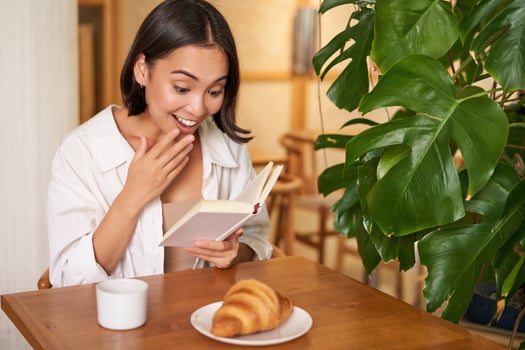 Asian girl looks amazed at pages, reading with excitement, sitting in cafe and eating croissant photo