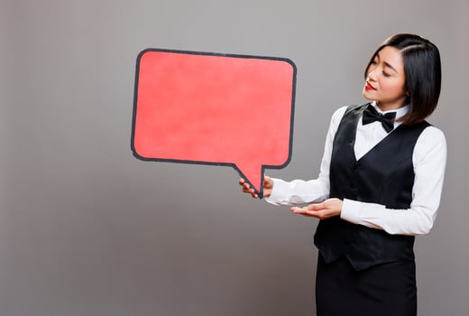 Young asian waitress holding blank chat balloon mockup while posing in studio. Attractive receptionist in professional uniform showing empty speak cloud with copy space for promo message photo
