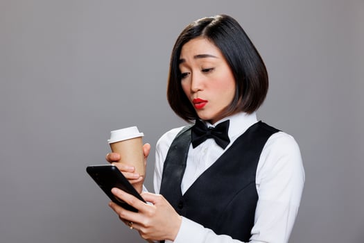 Asian woman in waitress uniform sending message on mobile phone during coffee break. Young receptionist talking on mobile phone social media and drinking hot beverage to go photo