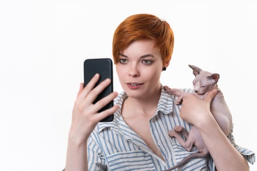 Redhead young woman hugging cat, having video-call holding smart phone in hand shooting selfie on camera. Studio shot on white background. Hipster in striped white-blue shirt. Part of series. photo