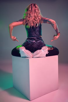 Young fitness blonde woman wearing blue velour booty shorts posing on camera, sits on top of big cube, back view photo