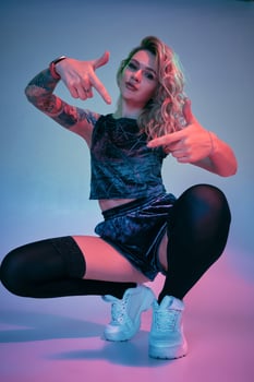 Young fitness blonde woman wearing blue velour booty shorts squats in front of camera, looking at camera, pink and blue background photo