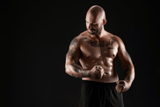Athletic bald, bearded, tattooed man in black shorts is posing against a black background. Close-up portrait. photo