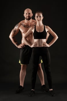 Athletic man in shorts and sneakers with brunette woman in leggings and top posing on black background. Fitness couple, gym concept. photo