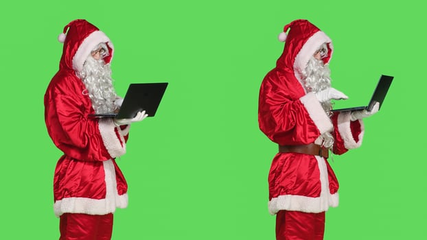 Santa claus man on online videocall waving at laptop screen to discuss with people about gifts, standing over greenscreen background. Person in red festive suit on teleconference call. photo