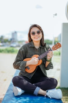 Playing Ukulele of Young Beautiful Asian Woman Wearing Jacket And Black Jeans Posing Outdoors photo