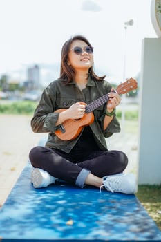 Playing Ukulele of Young Beautiful Asian Woman Wearing Jacket And Black Jeans Posing Outdoors photo