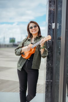 Playing Ukulele of Young Beautiful Asian Woman Wearing Jacket And Black Jeans Posing Outdoors photo