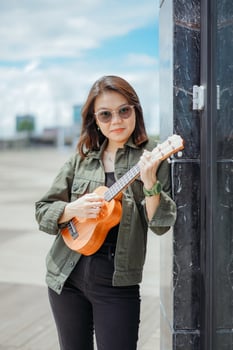 Playing Ukulele of Young Beautiful Asian Woman Wearing Jacket And Black Jeans Posing Outdoors photo