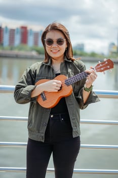 Playing Ukulele of Young Beautiful Asian Woman Wearing Jacket And Black Jeans Posing Outdoors photo