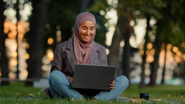 Happy smiling muslim islamic business woman girl female student wears hijab sitting on green grass lawn in city park using laptop for distant e-learning work chatting online checking mail with coffee photo