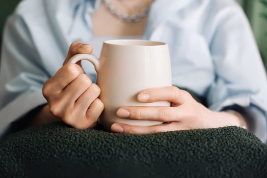 Woman Enjoying Tea on Living Room Couch, Cropped View. Cozy Domestic Scene. Young Woman Unwinding with Tea in Living Room Interior. Female hands holding a mug with tea or coffee. Close-up photo