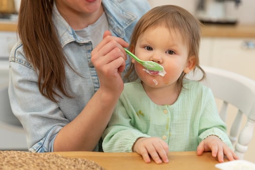 Happy family at home. Mother feeding her baby girl from spoon in kitchen. Little toddler child with messy funny face eats healthy food at home. Young woman mom giving food to kid daughter photo