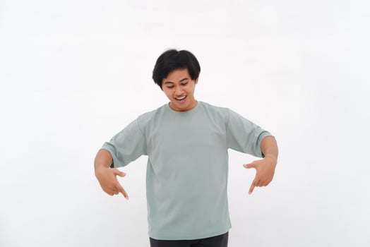 Excited Asian male employee wearing a T-shirt pointing at the copy space below him, isolated by a white background photo