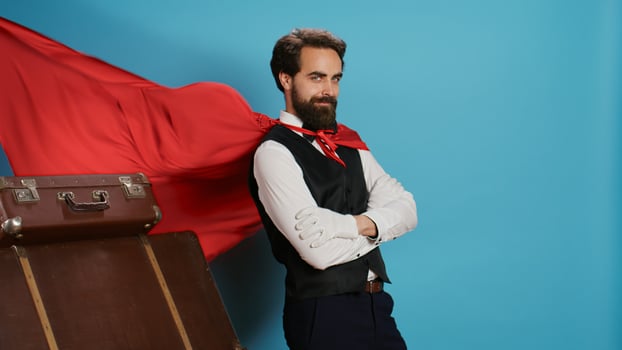 Hotel worker with red mantle to help guests with trolley bags, pretending to be fantasy cartoon superhero character on camera. Bellhop acting like comic superhuman with cape against background. photo
