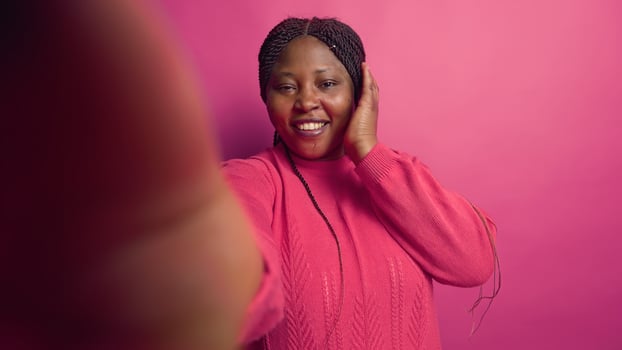 African american female vlogger talking on online video call with webcam, grasping smartphone. Enthusiastic black woman doing vlog content with digital mobile phone against vibrant pink background. photo