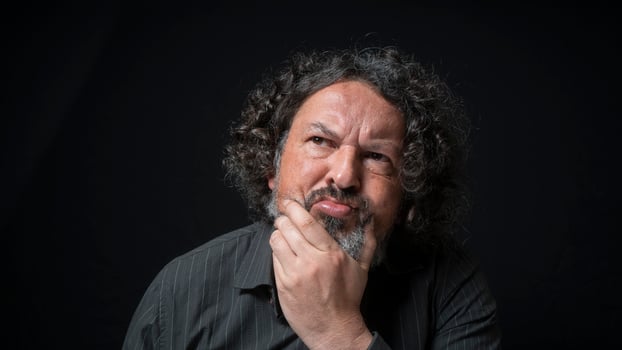 Man with white beard and black curly hair with indecisive expression, hand on chin, wearing black shirt against black background photo