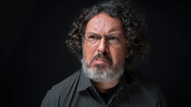 Man with white beard and black curly hair with frustrated expression, grimacing with mouth, wearing black shirt against black background photo